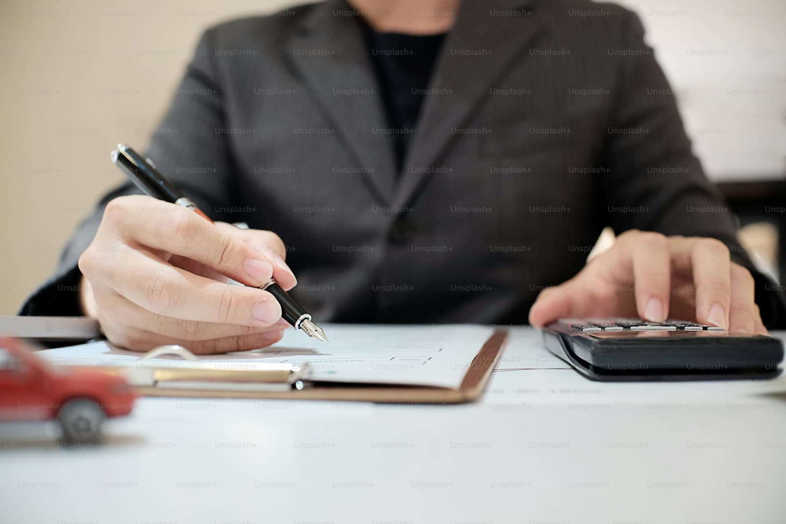 Person in a dark suit writing on paperwork while using a calculator at a desk
